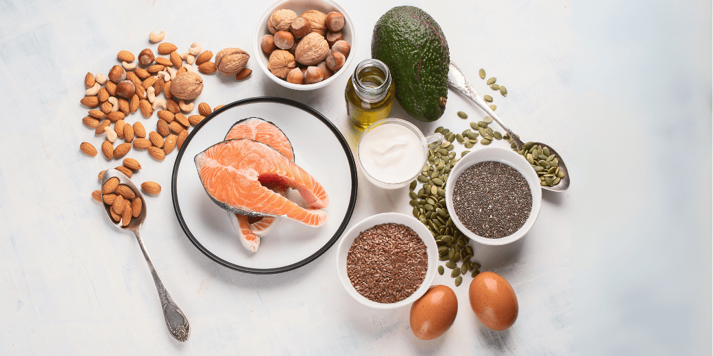 Verschiedene Omega 3 reiche Lebensmittel wie Lachs, Avocado, Nüsse, Samen, Eier und Öl liegen dekorativ auf einem hellen Untergrund