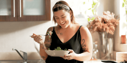 Lächelnde Frau mit Tattoos isst einen frischen Salat in ihrer Küche – Ausdruck für gesunde Ernährung, Selbstliebe und Lebensfreude.