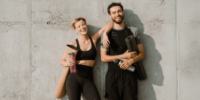 Lächelndes sportliches Paar in Trainingskleidung lehnt mit Yogamatten und Trinkflaschen an einer Betonwand.