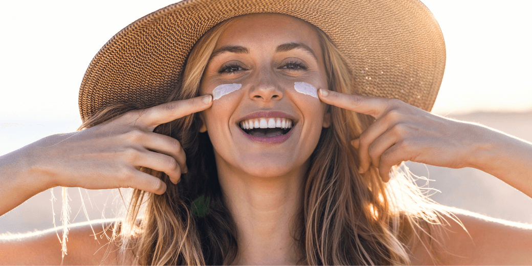 Lächelnde Frau mit Sonnenhut trägt Sonnencreme unter den Augen auf