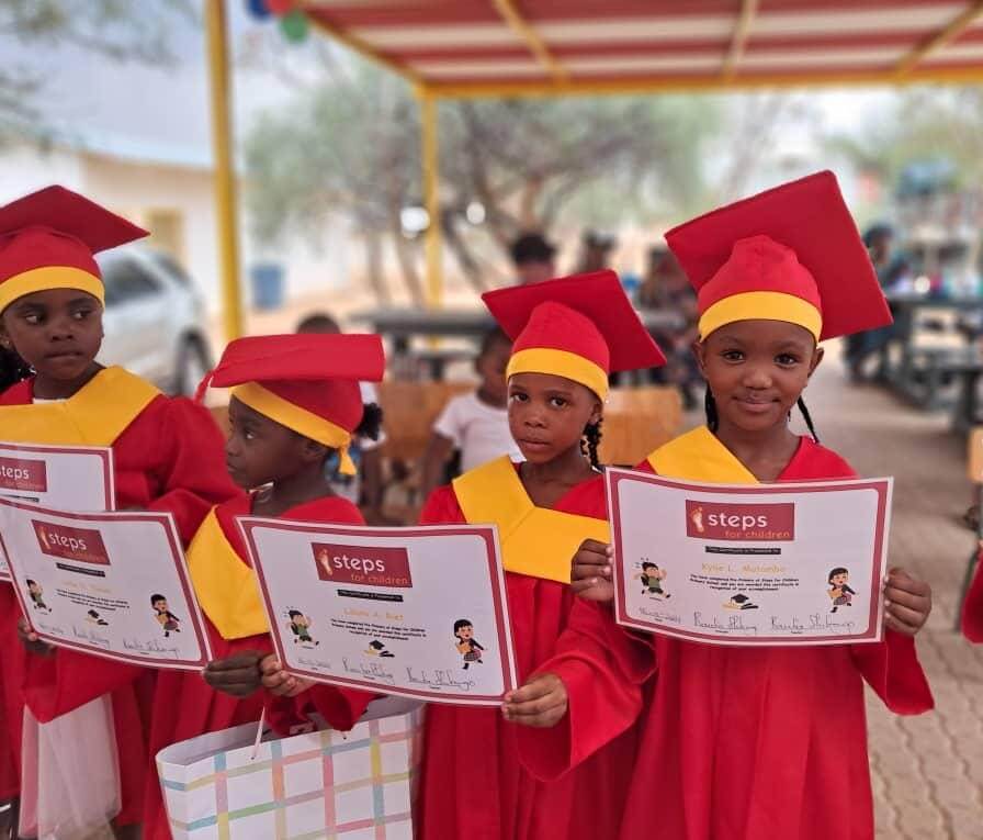 Vier Kinder in roten und gelben Abschlussgewändern halten stolz ihre steps-for-children Zertifikate in den Händen. Sie stehen nebeneinander und lächeln – ein Moment voller Freude, Bildung und Zukunftshoffnung.