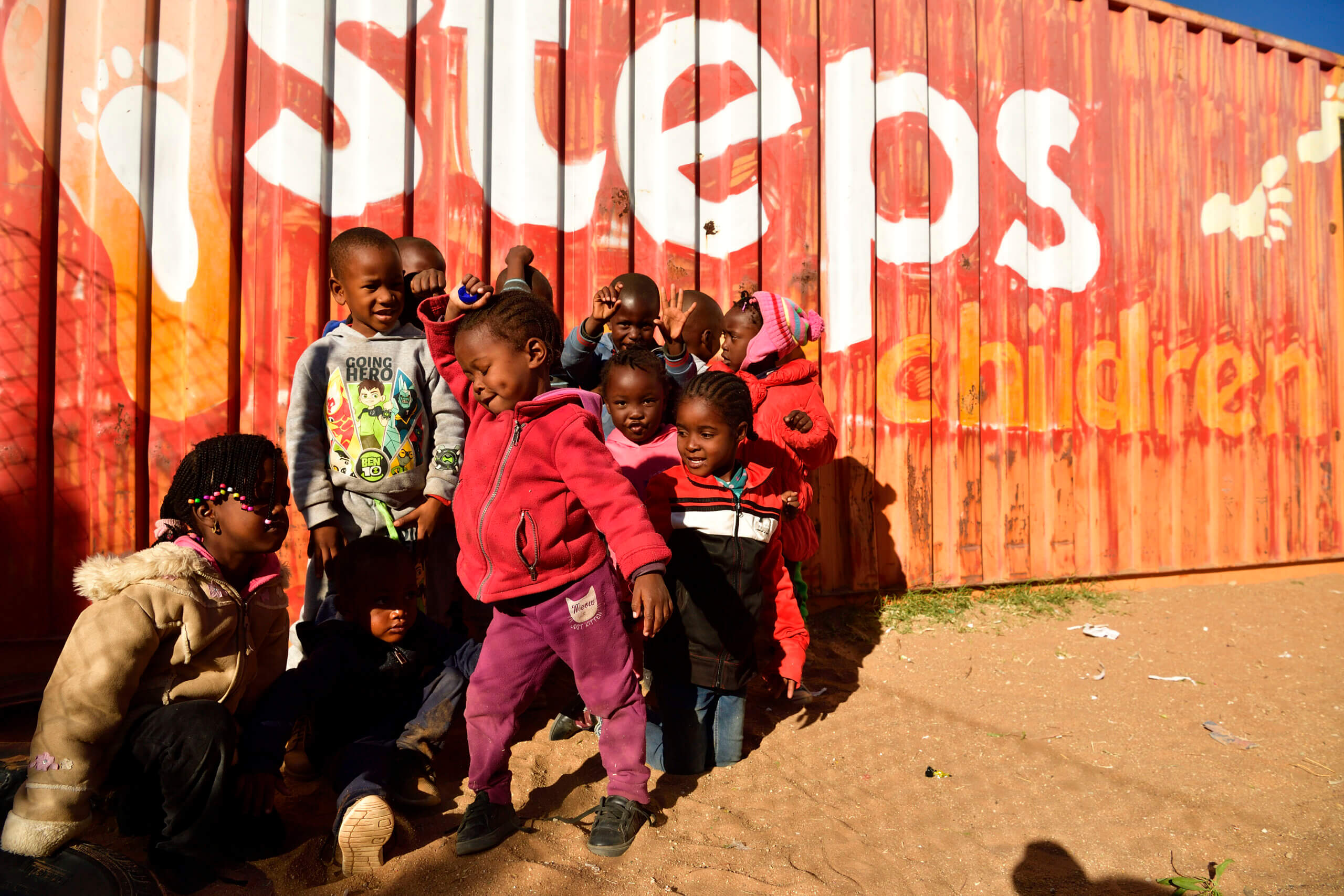 Gruppe lachender Kinder spielt vor einem orangefarbenen Container mit der Aufschrift „steps for children“ – Symbol für Bildung, Gemeinschaft und Hoffnung.