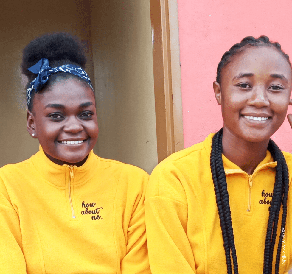 Drei junge Frauen sitzen nebeneinander und lächeln in die Kamera. Sie tragen gelbe Pullover und wirken fröhlich und zuversichtlich – ein Moment, der Gemeinschaft, Bildung und Zukunftschancen symbolisiert.