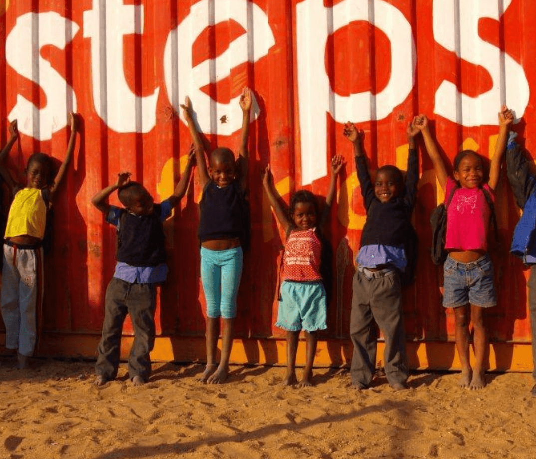 Gruppe lachender Kinder spielt vor einem orangefarbenen Container mit der Aufschrift „steps for children“ – Symbol für Bildung, Gemeinschaft und Hoffnung.