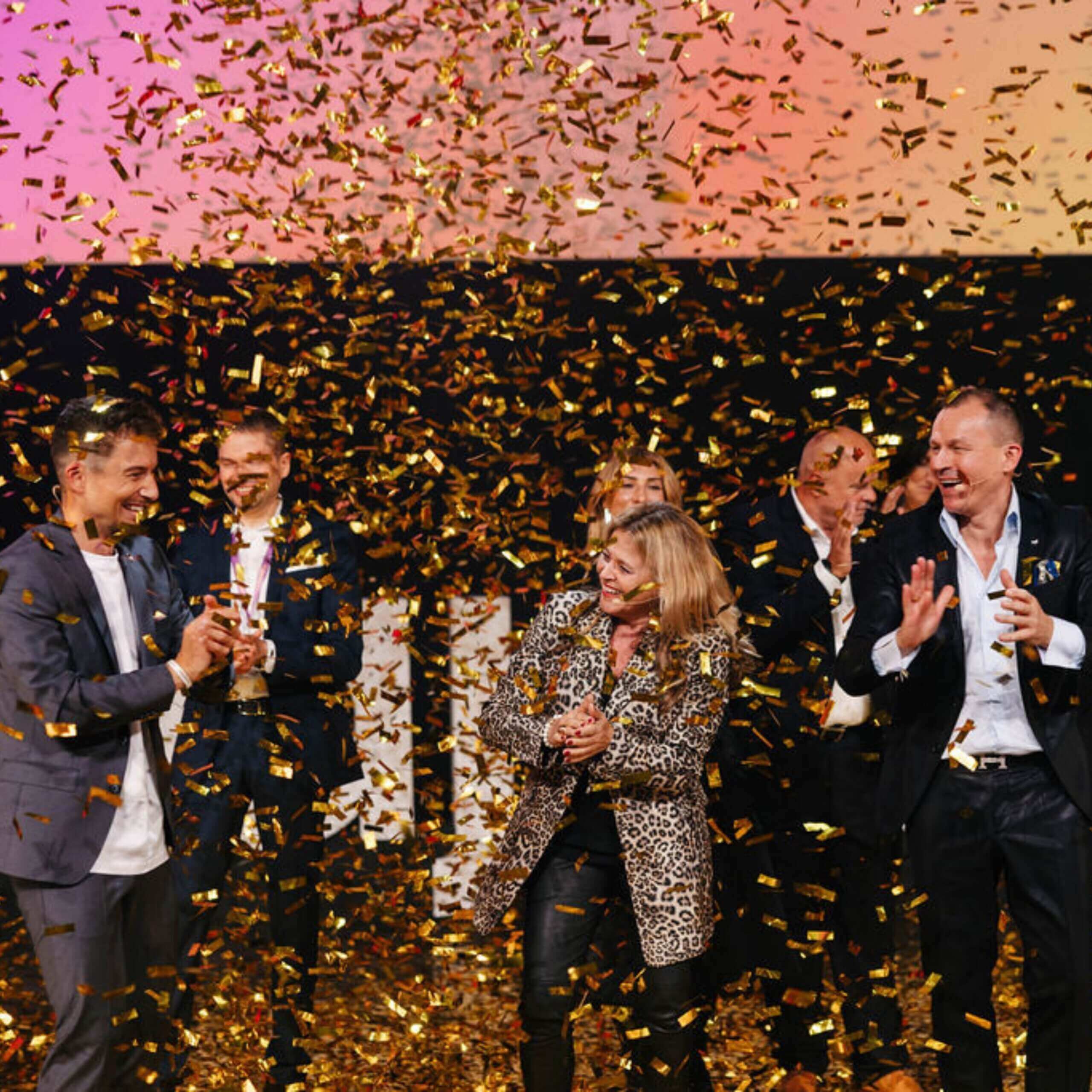 Auf der Bühne stehen mehrere Menschen und feiern gemeinsam. Goldene Konfetti regnen auf sie herab. Die Menschen klatschen, lachen und wirken fröhlich. Im Hintergrund ist ein rosa-orangefarbener Farbverlauf zu sehen.